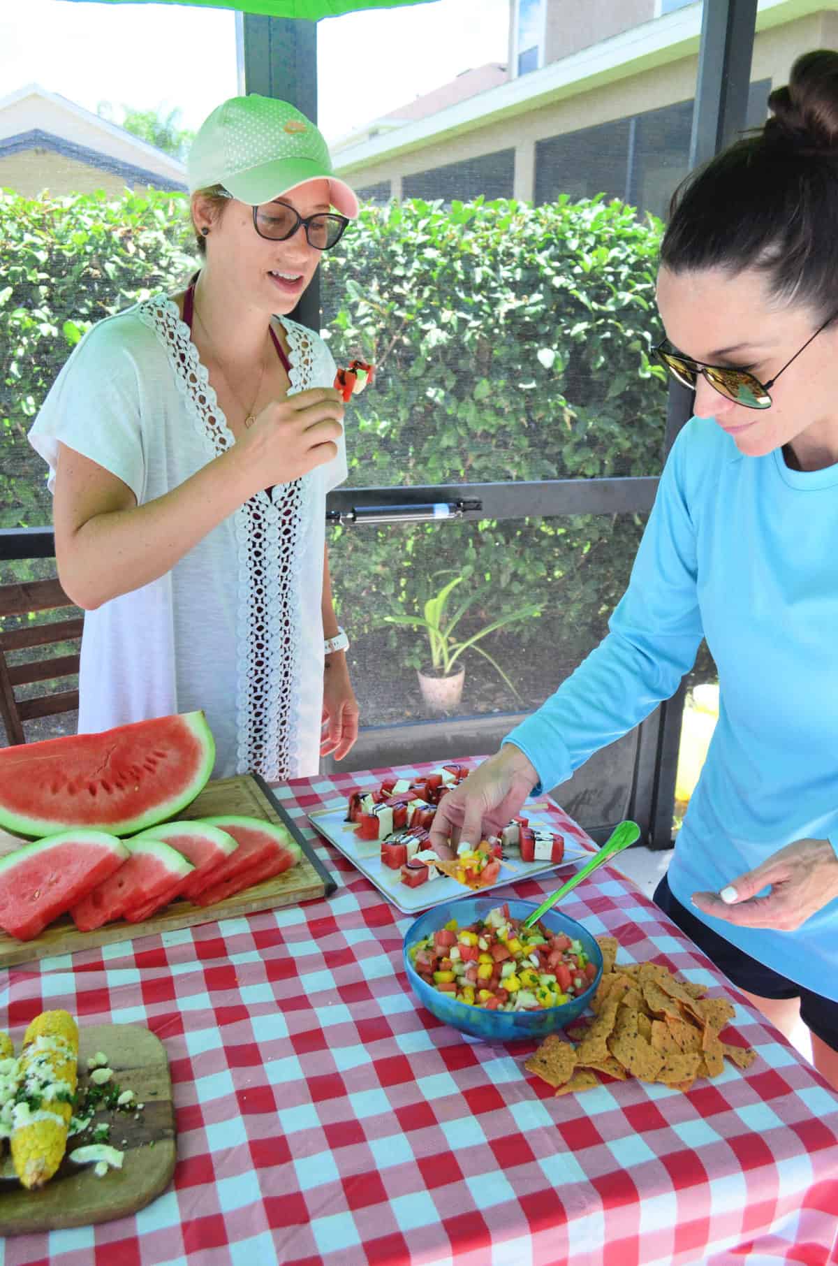 two women enjoying watermelon salsa at picnic table that also has watermelon feta skewers and slices.