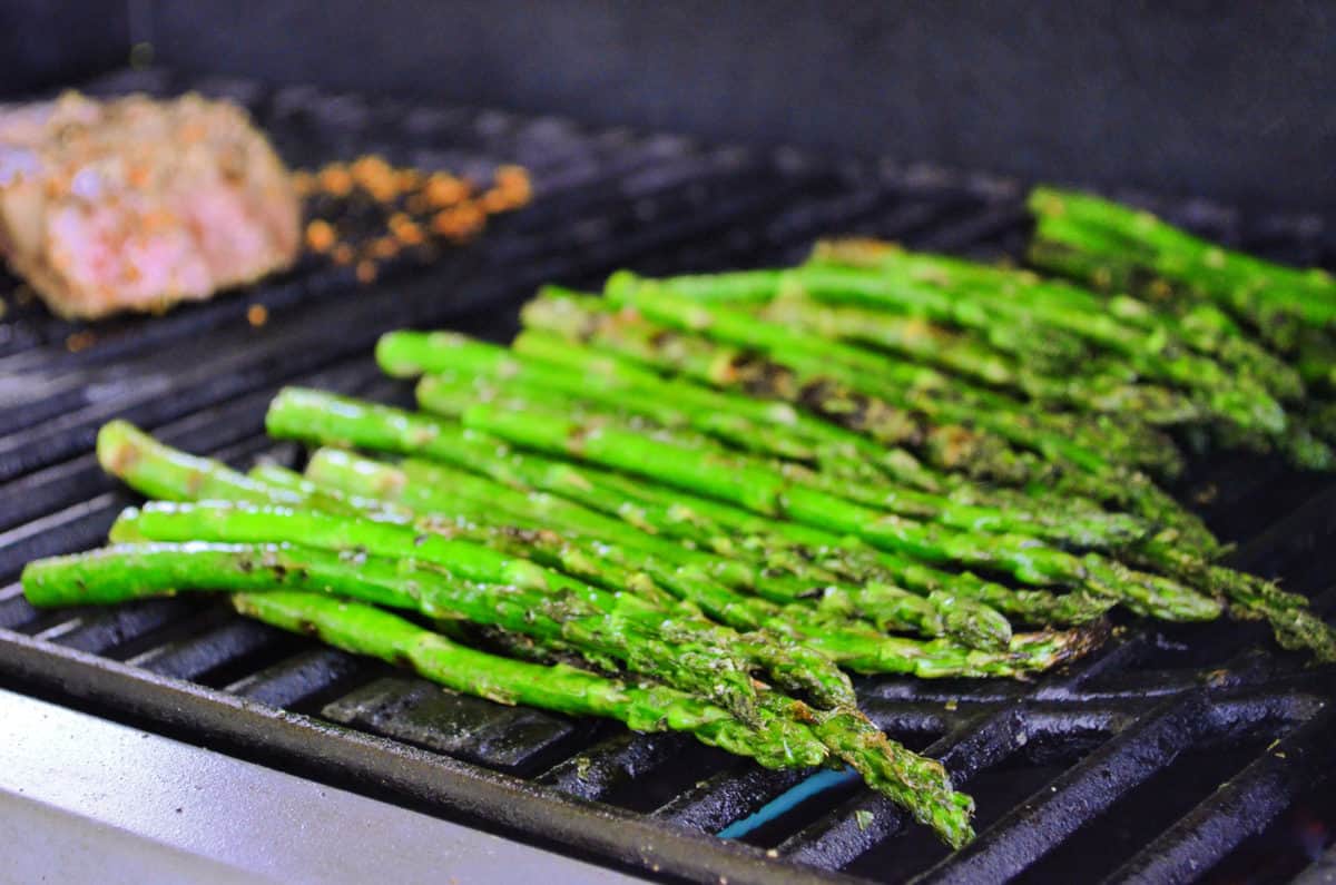 Side view of Italian Herb Asparagus on grill with meat blurred in background.