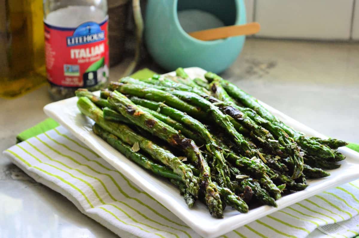 grilled asparagus on rectangular platter with bottle of italian herbs blurred in background.