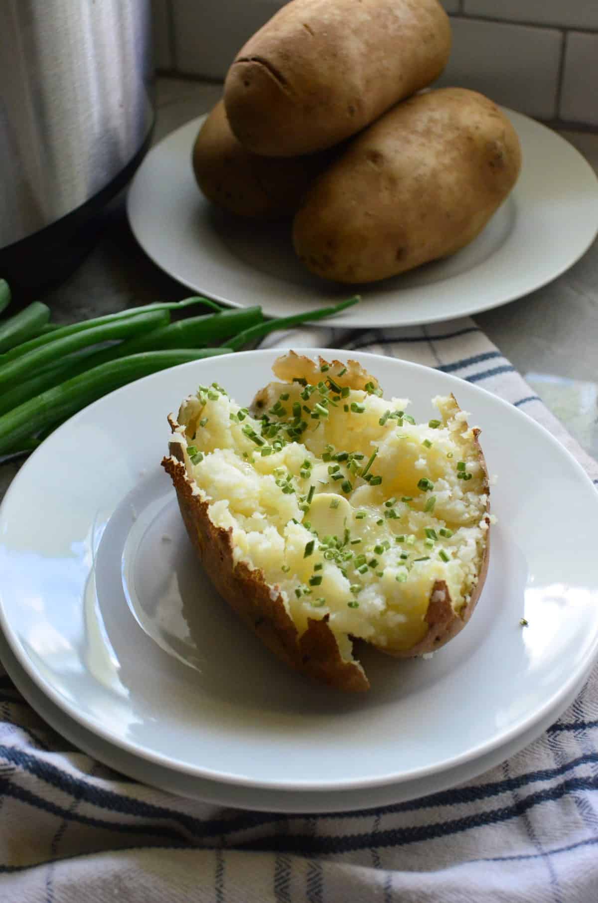 Plated halved baked potato topped with butter and chives with whole potatoes in background.