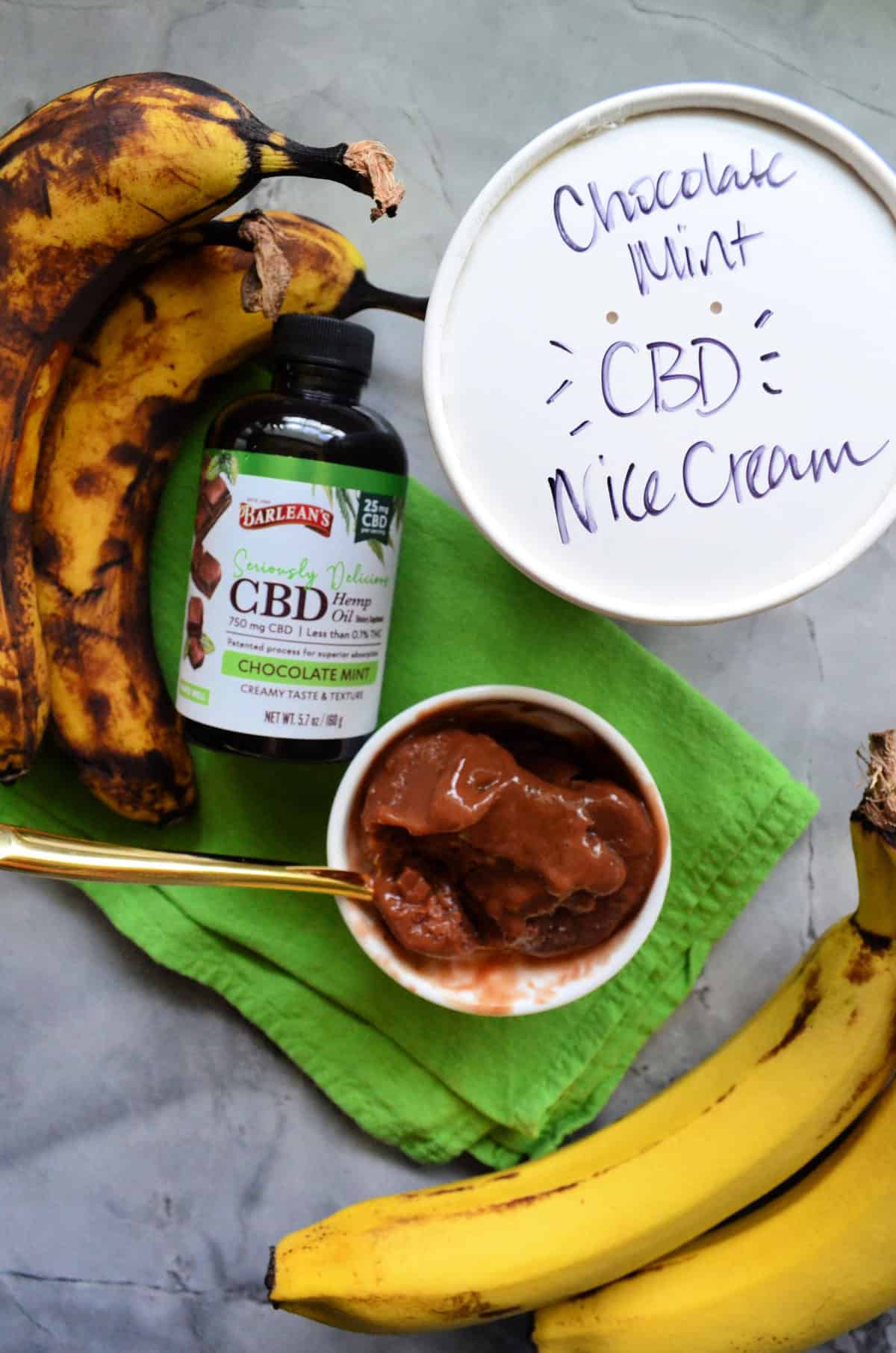 top view bowl of chocolate ice cream with spoon next to bananas, CBD oil bottle, and labeled pint.