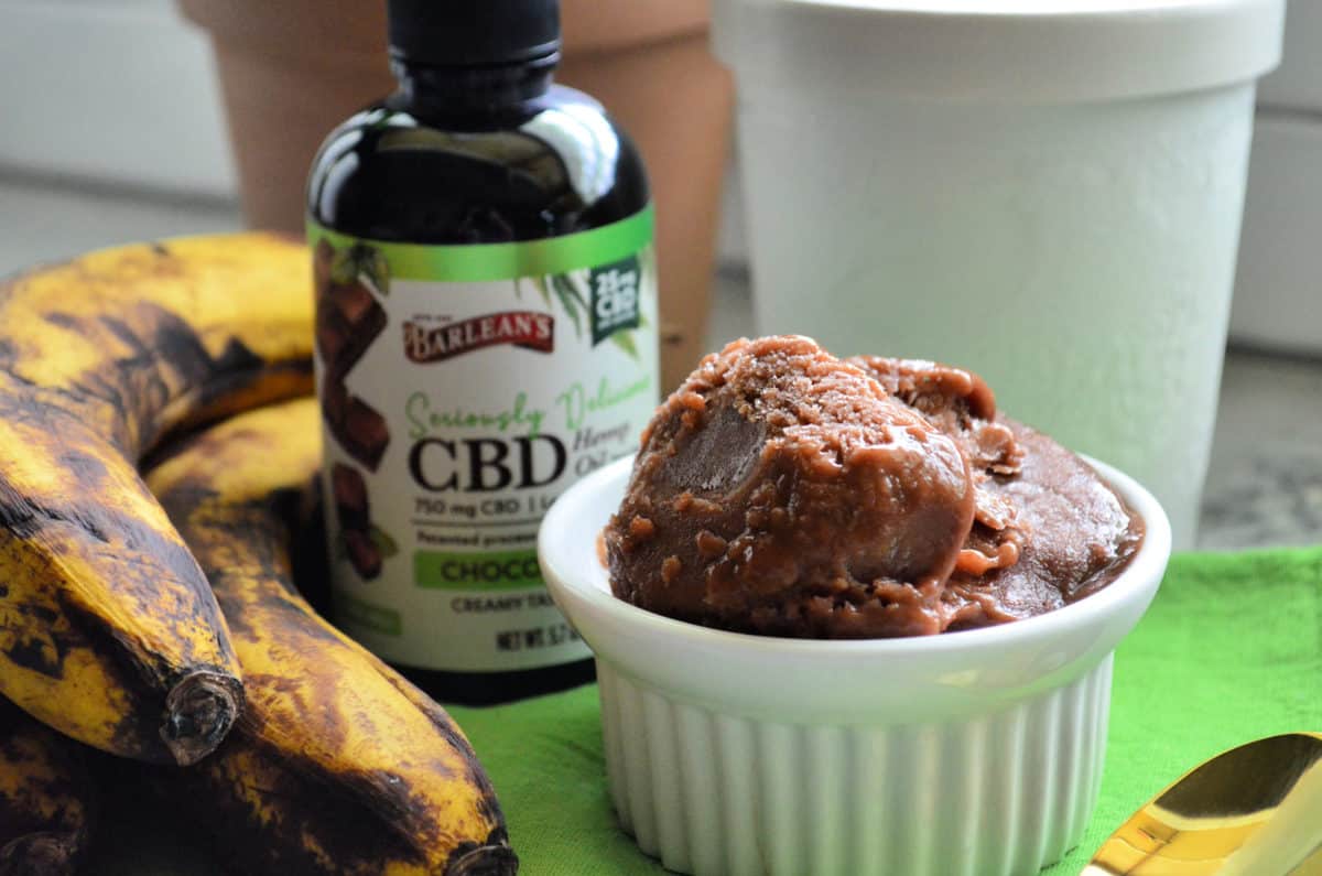 side view bowl of chocolate ice cream with spoon next to bananas, CBD oil bottle, and labeled pint.