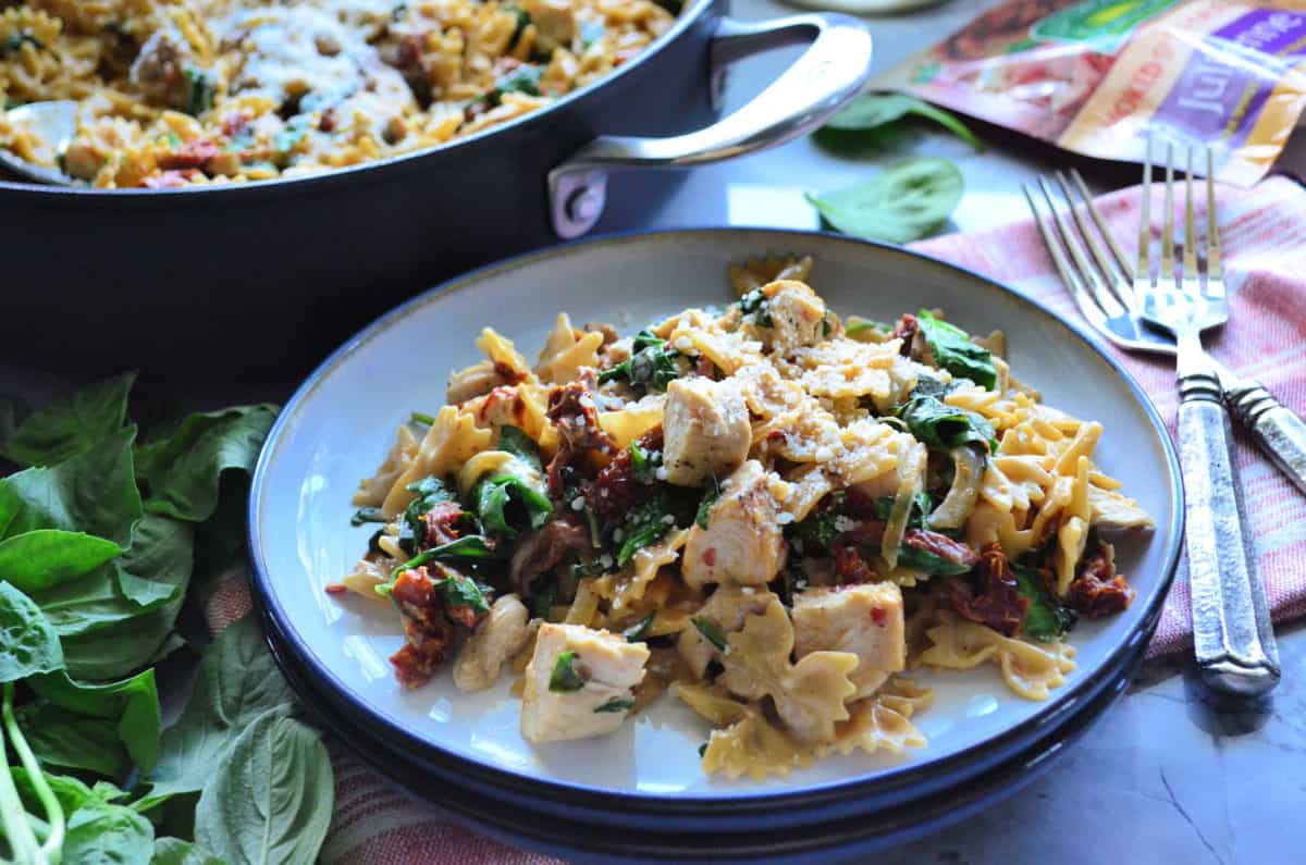 bowtie pasta, basil, sundried tomato chicken, and parmesan on plate near forks, in front of skillet.