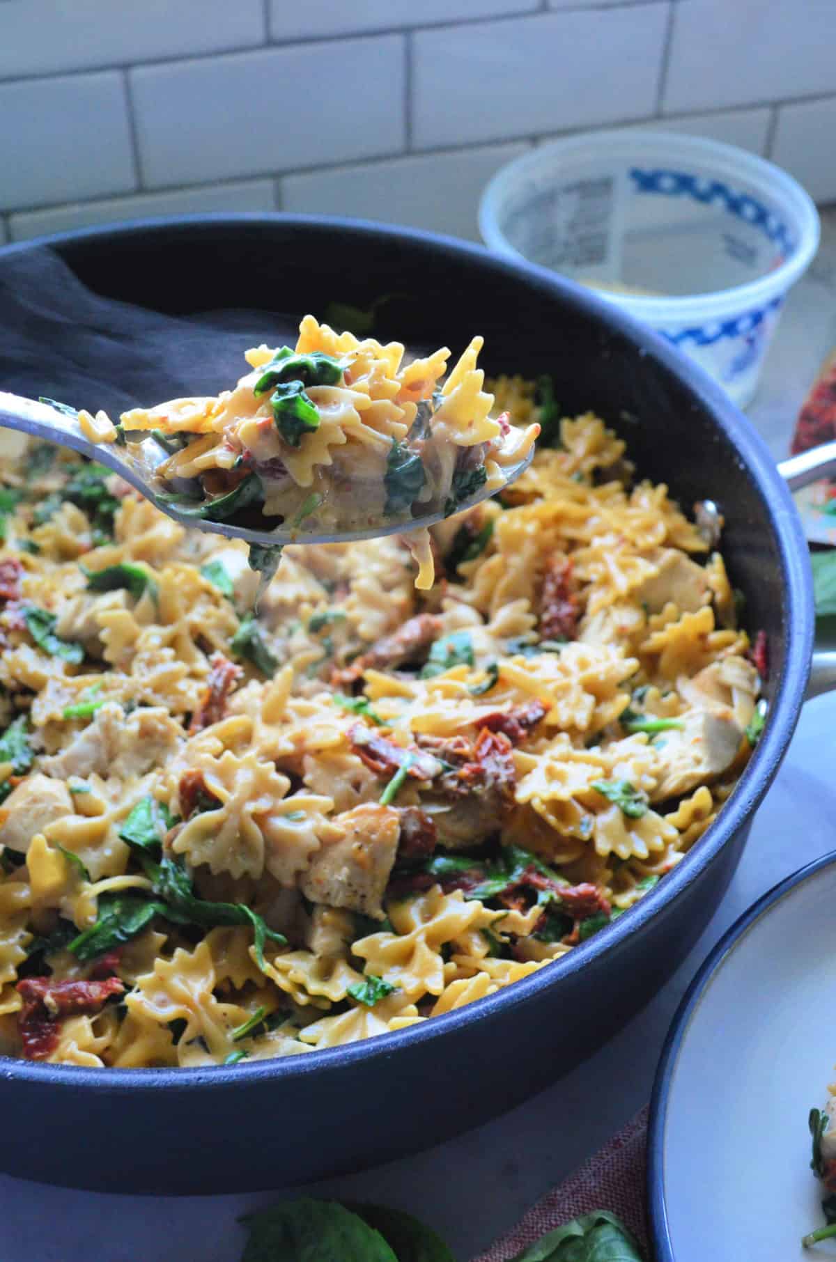 fork of bowtie pasta, basil, sundried tomato chicken, and parmesan held over skillet.