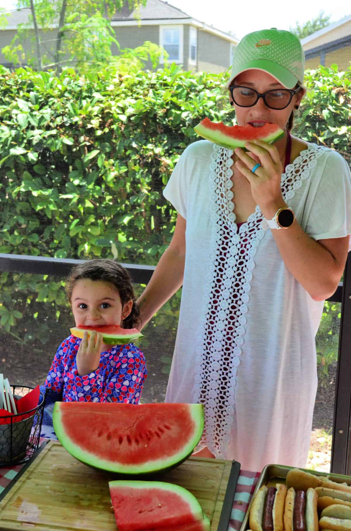 Mother and daughter standing next to eachother, each enjoying a slice of watermelon at picnic table.