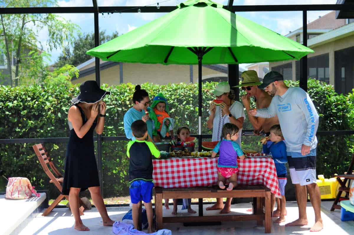 5 adults and 5 kids enjoying snacks around picnic table outside under green umbrella.