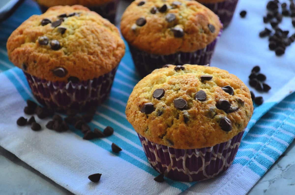 3 golden brown chocolate chip muffins on blue striped cloth scattered with chocolate chips.