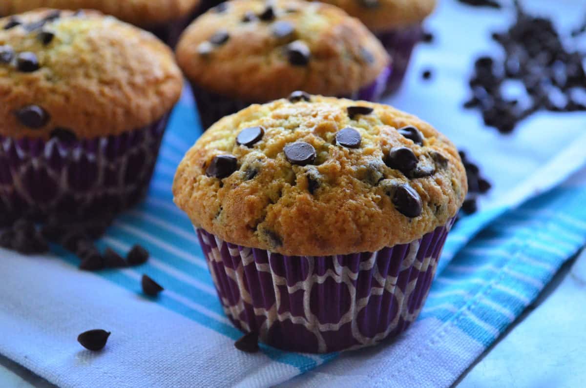 side view chocolate chip muffin on blue cloth scattered with chocolate chips and muffins in background.