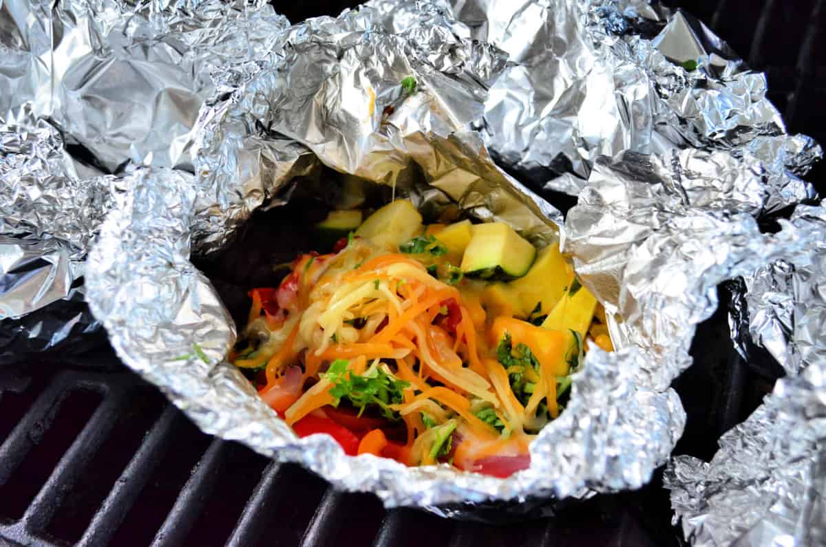 Aluminum foil packet on grill opened to show melting cheese with vegetables.