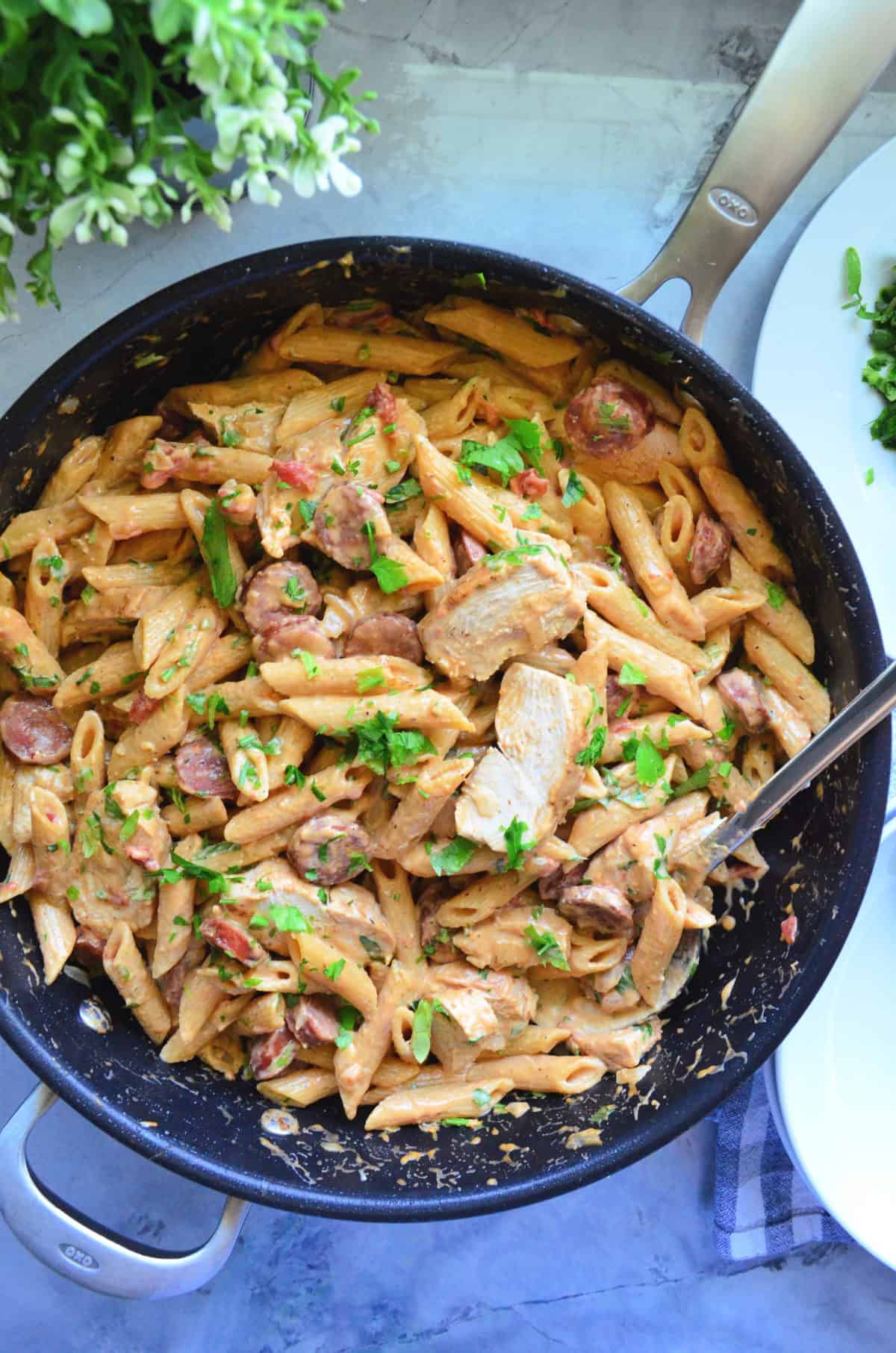 Top view of sliced sausage, penne, green herbs, chicken tossed in light sauce in pan with spoon.