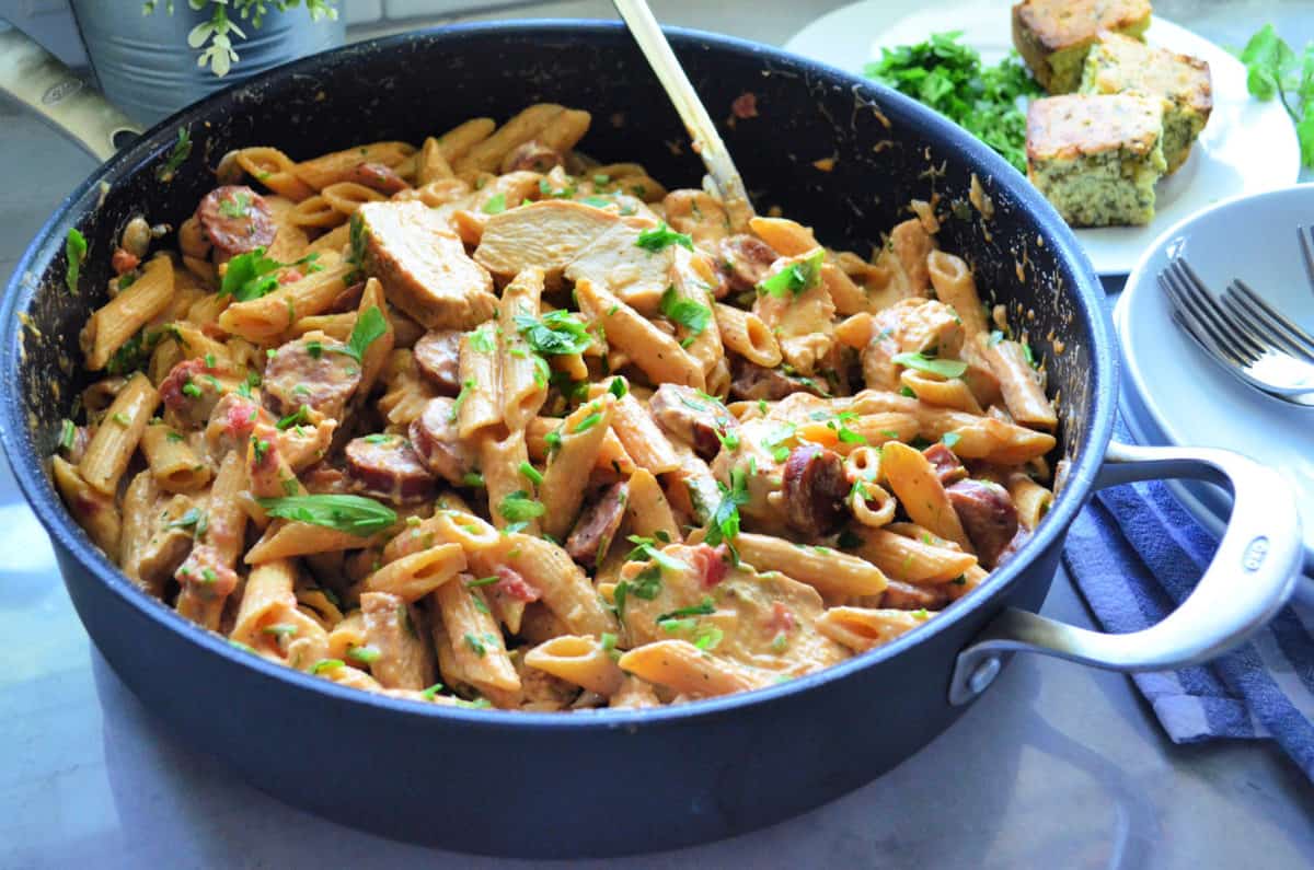 Top view of sliced sausage, penne, green herbs, chicken tossed in light sauce in pan.