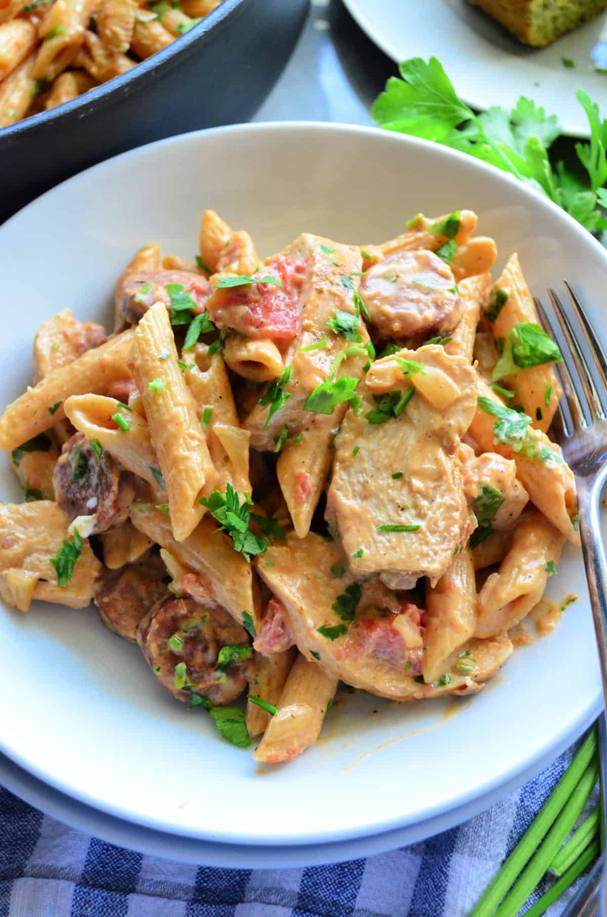 close up of of sliced sausage, penne, green herbs, chicken tossed in light sauce on plate with fork.