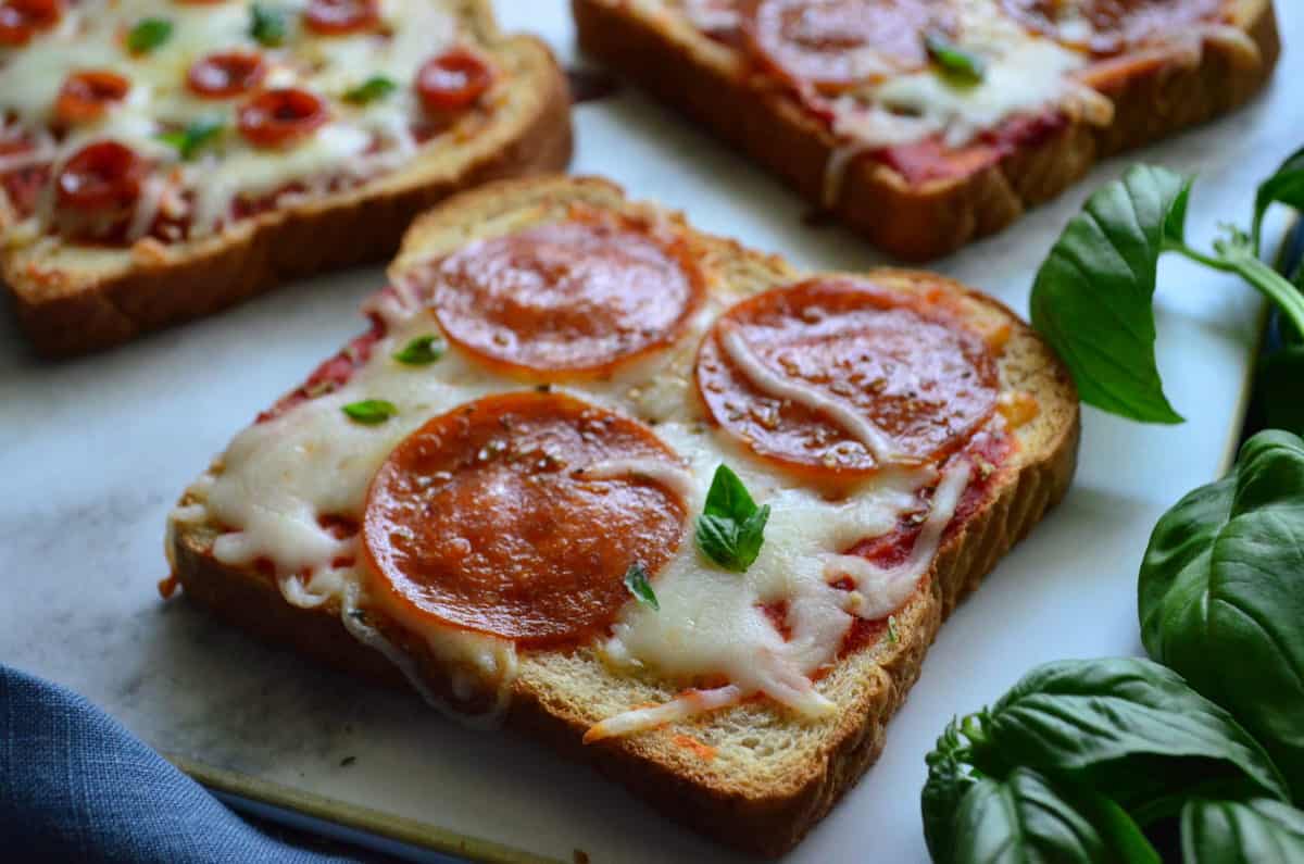 3 pieces of toast with marinara, pepperoni, and cheese on countertop next to basil.