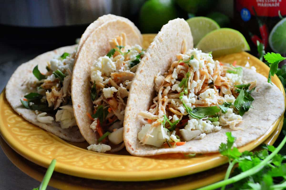 3 shredded chicken tacos served with lime on yellow pate next to blurred cilantro.
