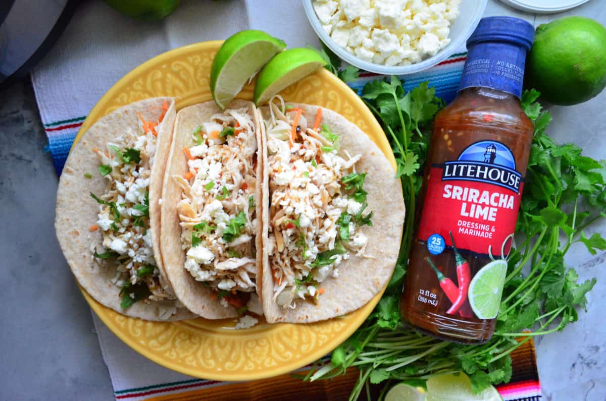 3 tacos filled with shredded chicken, cheese, and herbs near limes, cilantro, and litehouse cilantro lime dressing bottle.