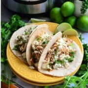 3 flour tortilla tacos filled with shredded chicken, cheese, and herbs surrounded by limes and cilantro.