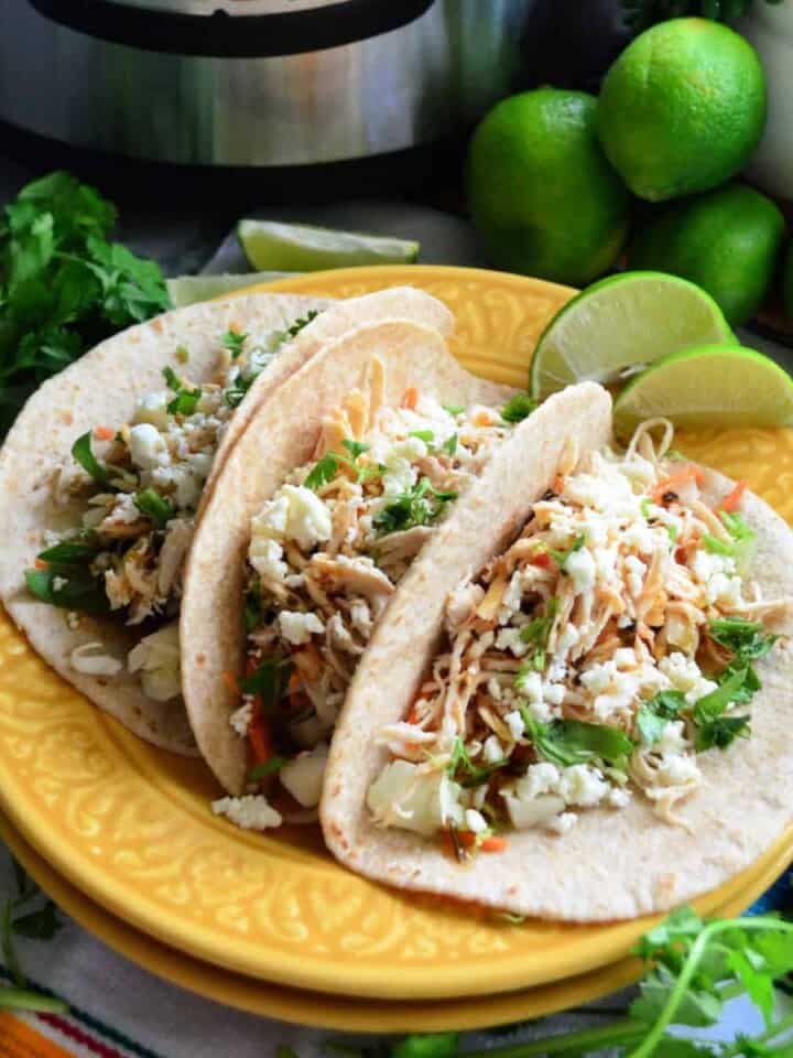 3 flour tortilla tacos filled with shredded chicken, cheese, and herbs surrounded by limes and cilantro.
