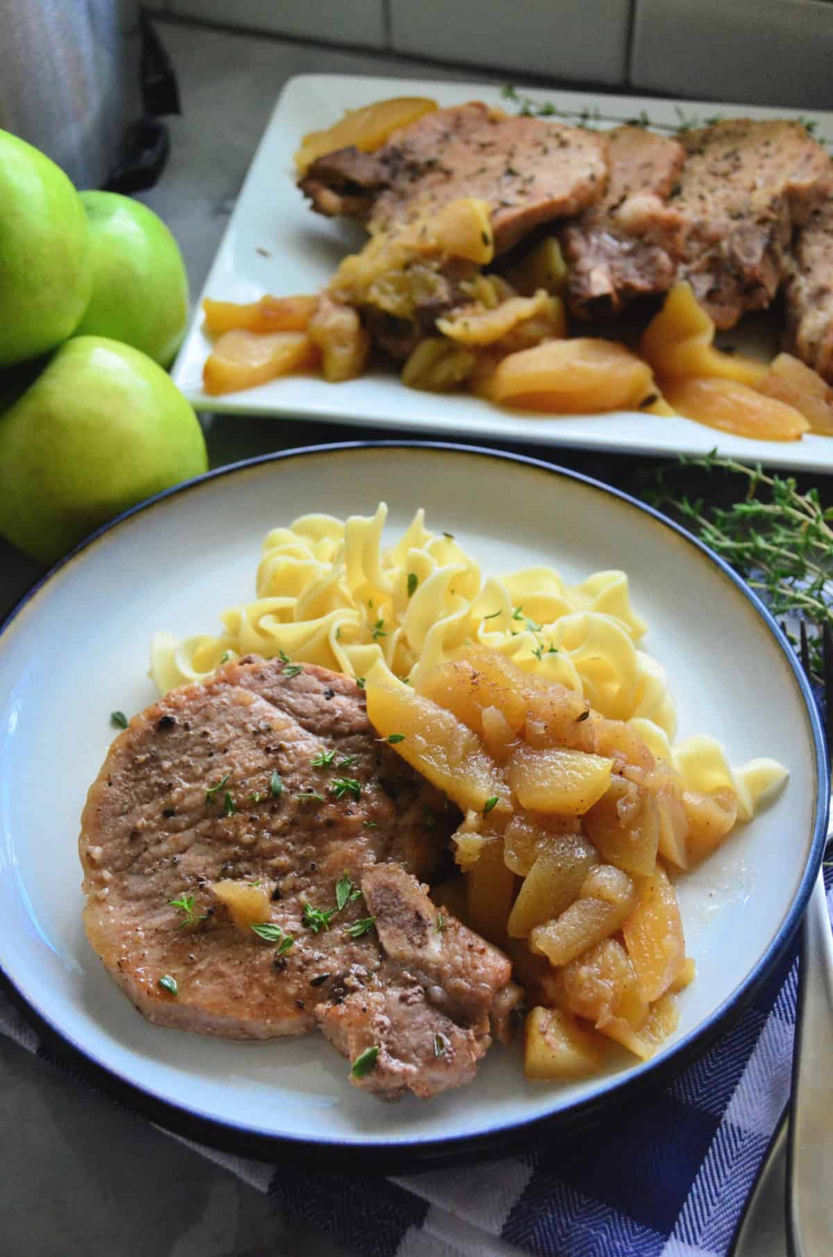 Plated sliced pork chops with cooked apples, egg noodles, and platter of pork chops in background.