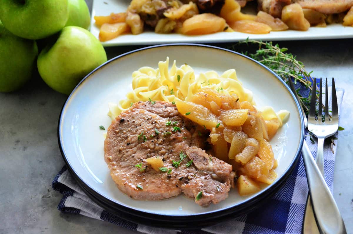 plated sliced pork chops with thyme , apples, and egg noodles on placemat with fork.