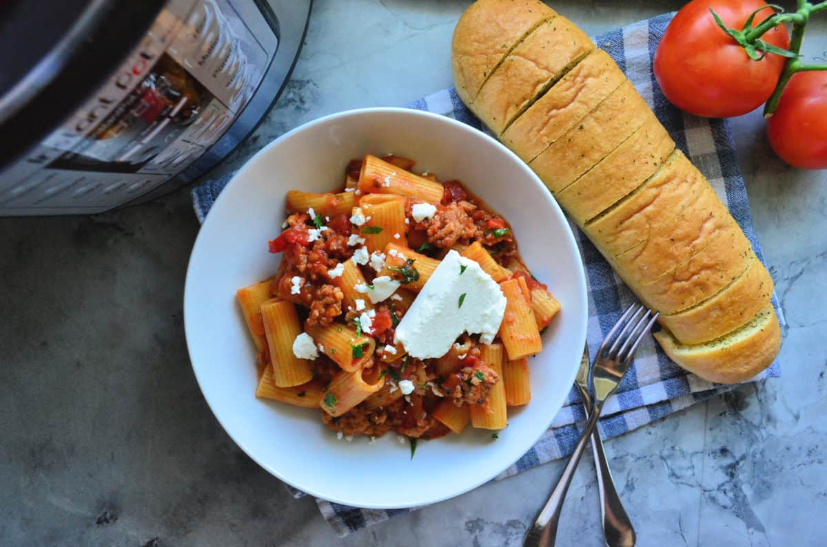 Plated Rigatoni noodles with meaty red sauce, basil, and cheese next to sliced baguette and tomatoes.