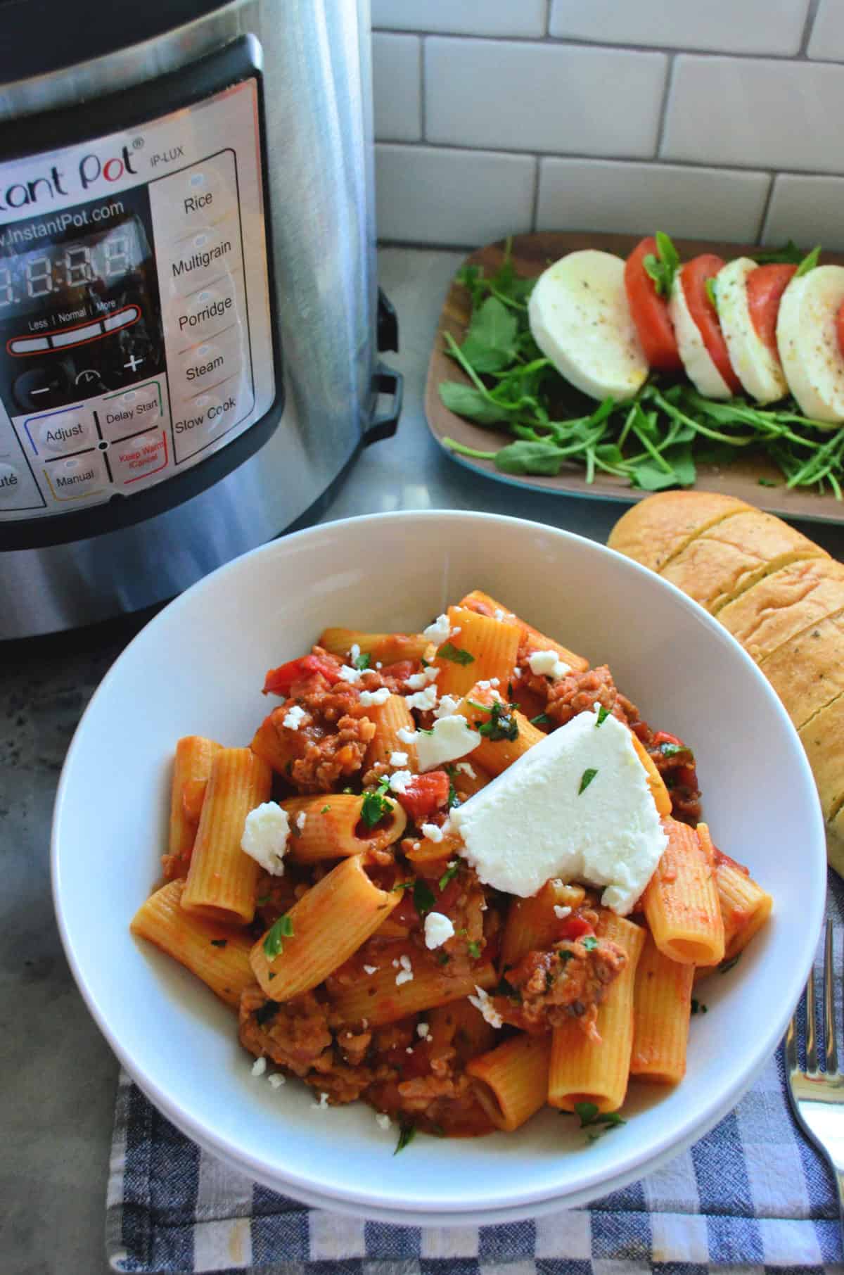 Plated Rigatoni noodles with meaty red sauce, basil, and cheese in front of instant pot and caprese platter.