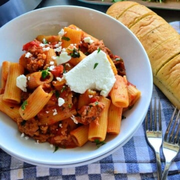 Bowl Rigatoni in meaty red sauce with goat cheese on placemat with forks and french bread.