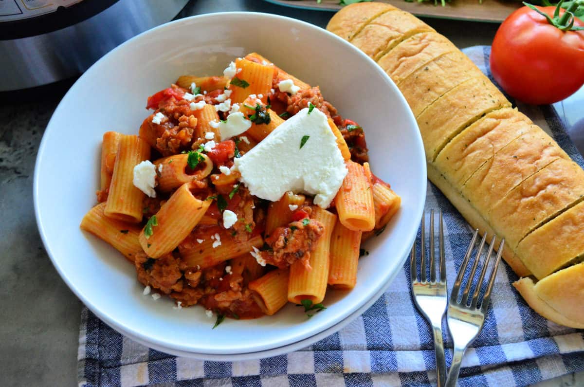Bowl Rigatoni in meaty red sauce with goat cheese on placemat with forks and french bread.