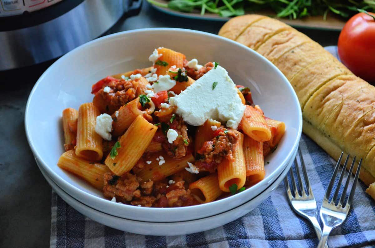 Plated Rigatoni noodles with meaty red sauce, basil, and cheese in front of baguette and tomato.