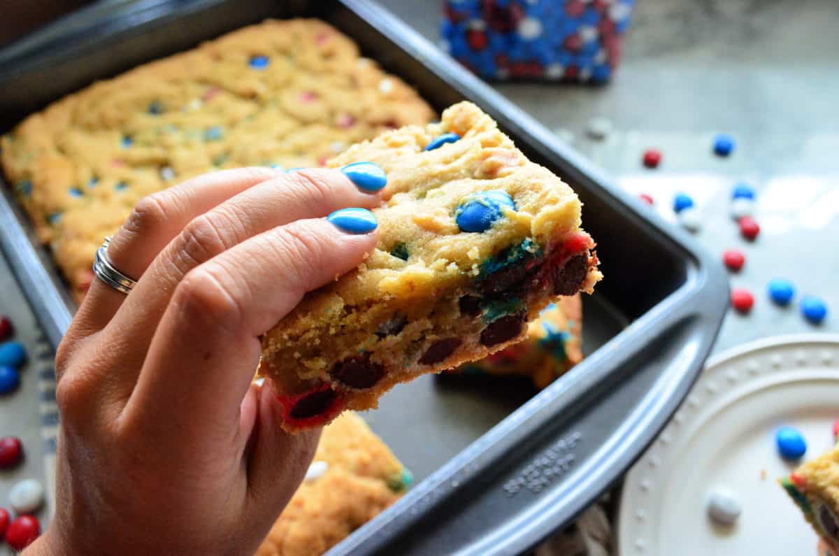 Hand with blue fingernails holding a cookie bar over pan of remaining cookie.