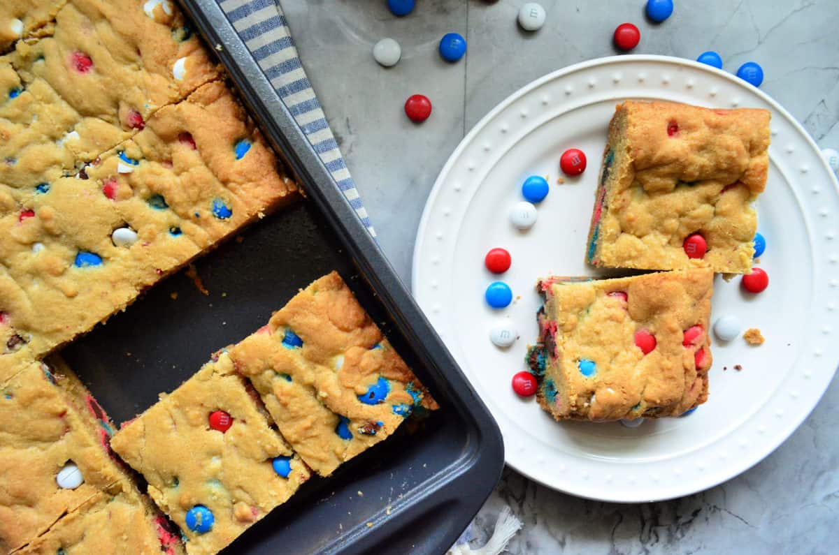 A pan of cookie bars with a white plate and 2 cookie bars right next to it.