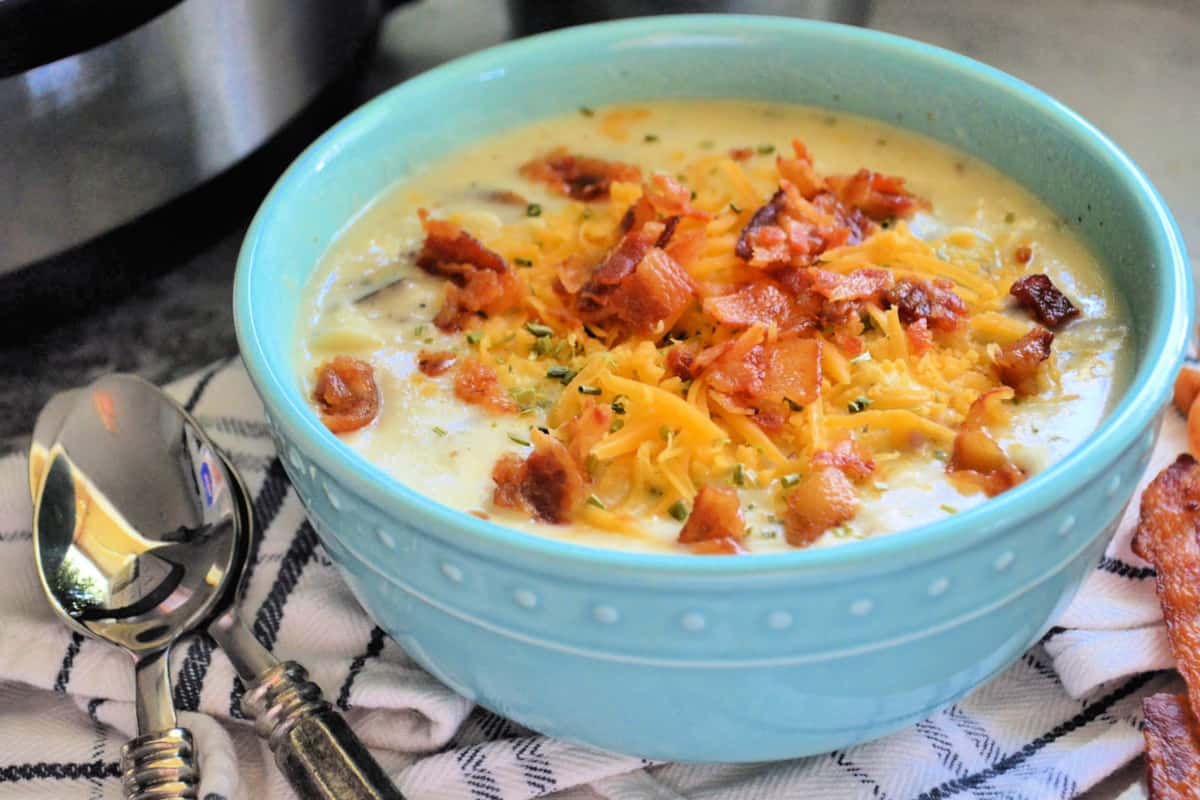 Bowl of creamy soup topped with herbs, bacon, and cheddar on tablecloth with spoons.