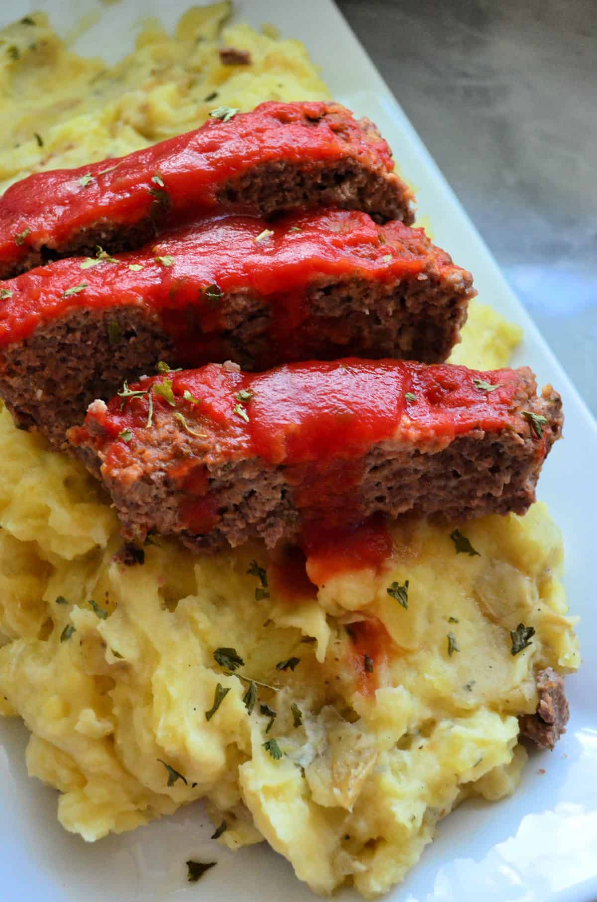 closeup of sliced meatloaf topped with herbs and red sauce over mashed potatoes.