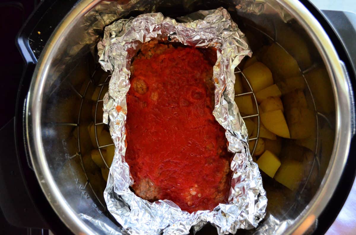 top view of meatloaf in tinfoil resting on wire stand over chopped potatoes.