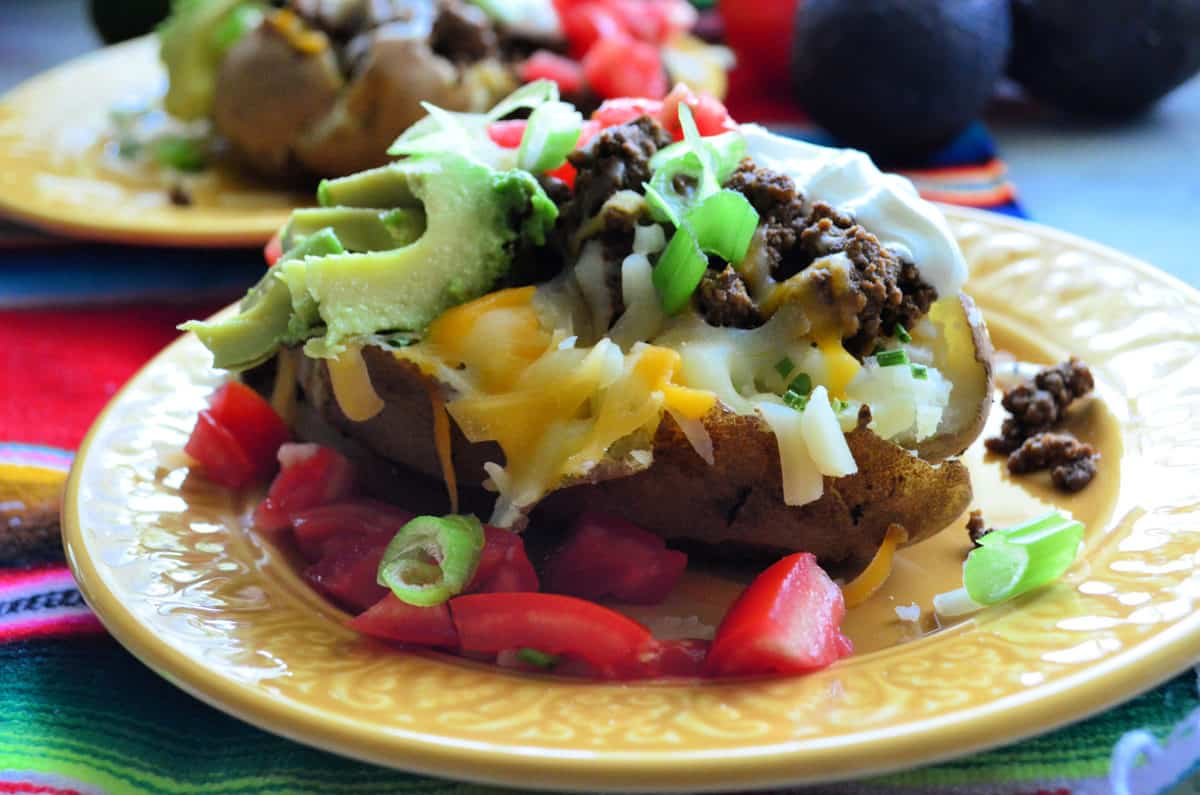 Side View Plated Baked potato loaded with sour cream, tomato, olives, avocado, and shredded cheese.