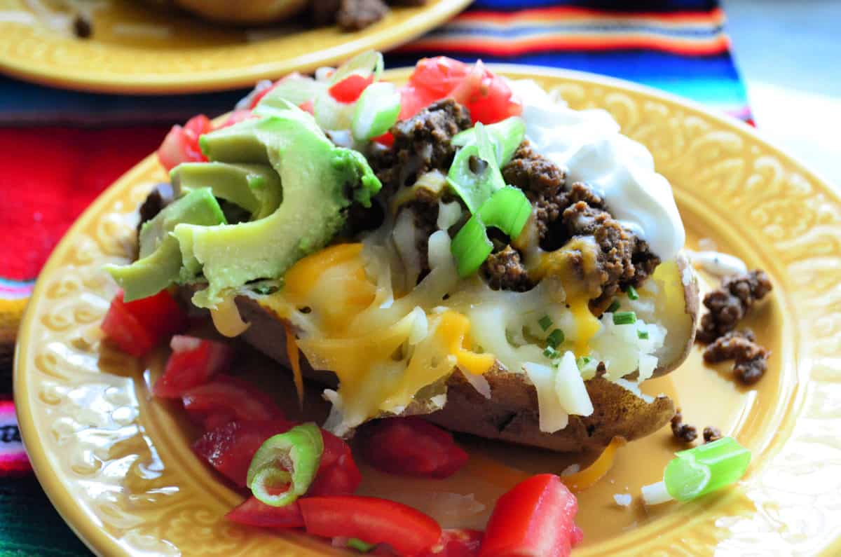split baked potato with sour cream, meat, tomato, cheese, avocado, green onions on yellow plate.