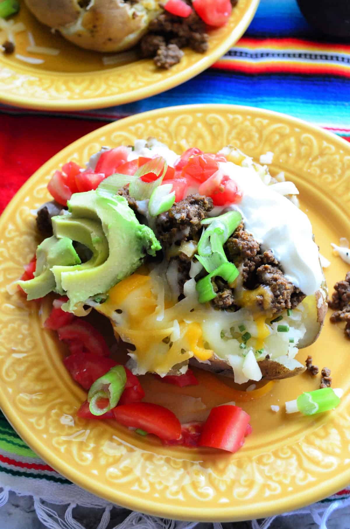 Top view split baked potato with sour cream, meat, tomato, cheese, avocado, green onions on plate.