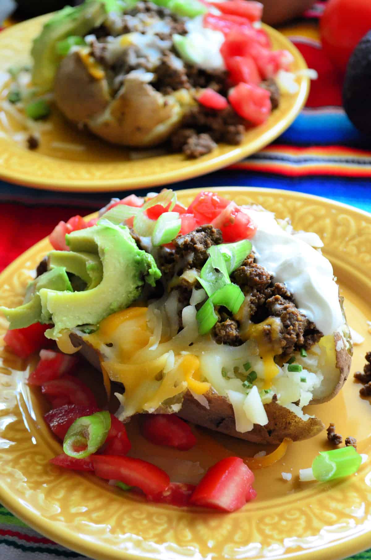 2 split baked potatoes with sour cream, meat, tomato, cheese, avocado, green onions on plates.