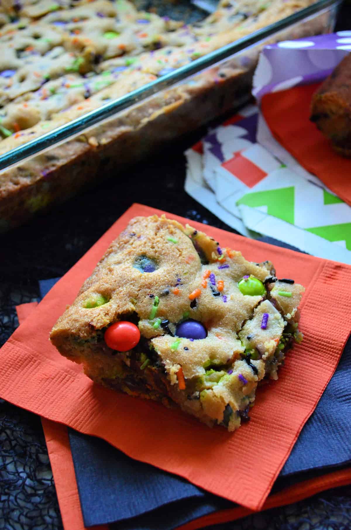 cookie bar square made with orange, purple, and green M&Ms and sprinkles on orange napkin.