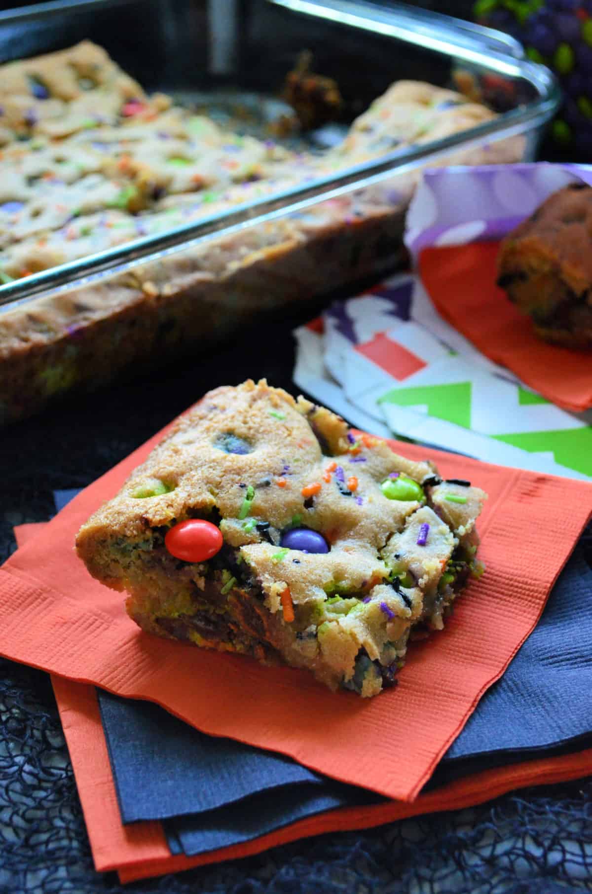 cookie bar square made with orange, purple, and green M&Ms and sprinkles on orange napkin.