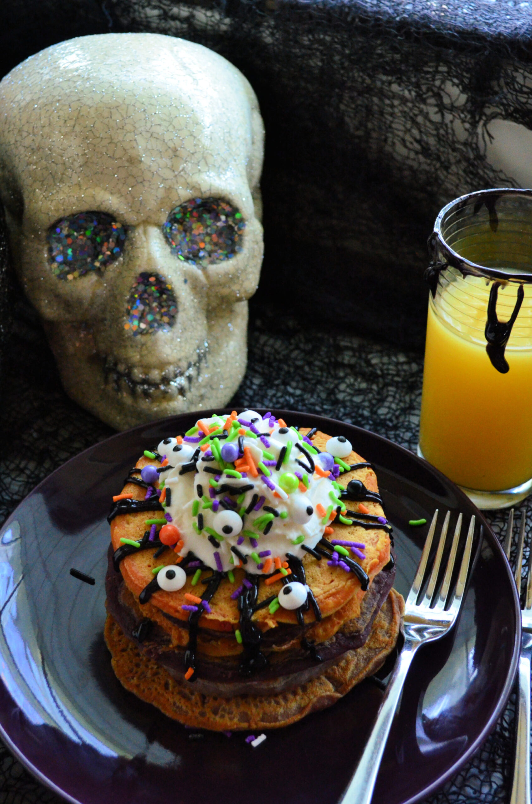 Orange colored pancakes topped with halloween themed sprinkles, googly eyes, whipped cream, and chocolate drizzles.