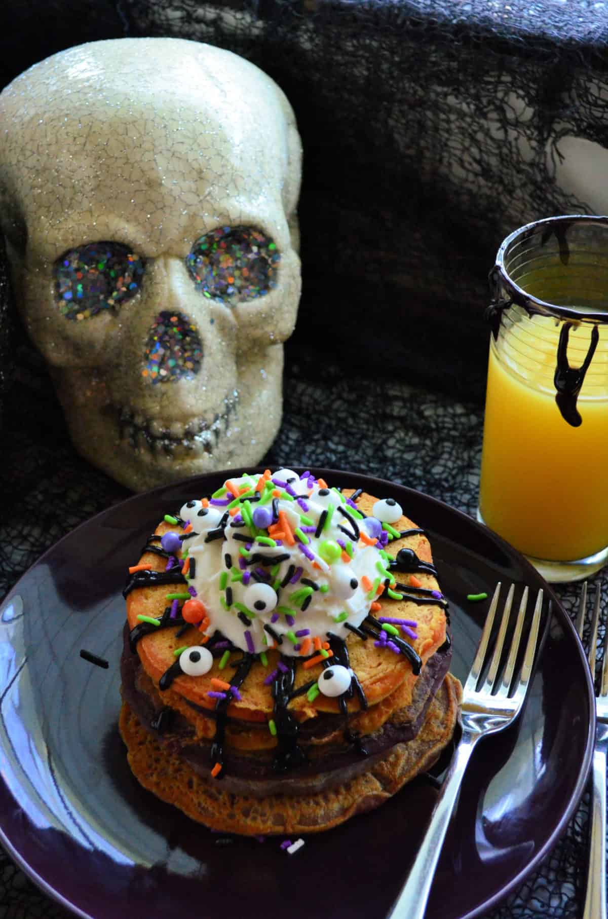 Orange pancakes stacked on black plate topped with candy eyes, whipped cream, and halloween sprinkles.