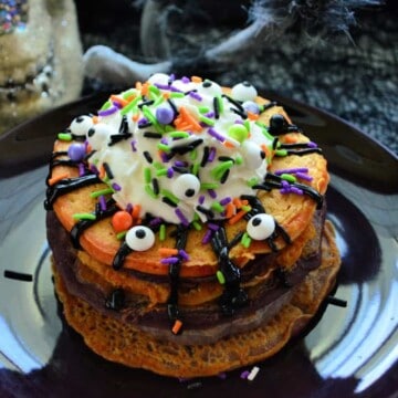 Orange pancakes stacked on black plate topped with candy eyes, whipped cream, and halloween sprinkles.