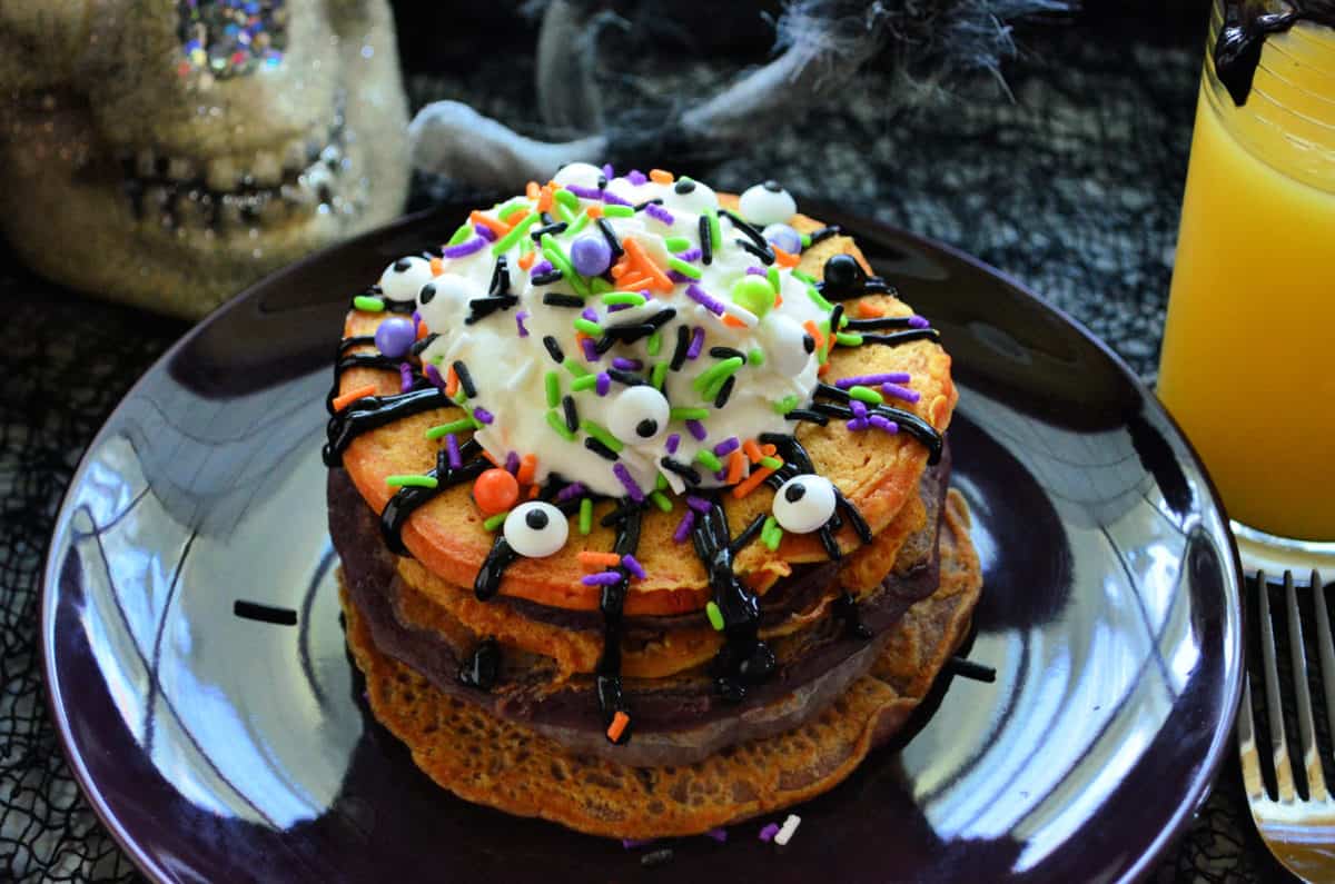 Orange pancakes stacked on black plate topped with candy eyes, whipped cream, and halloween sprinkles.