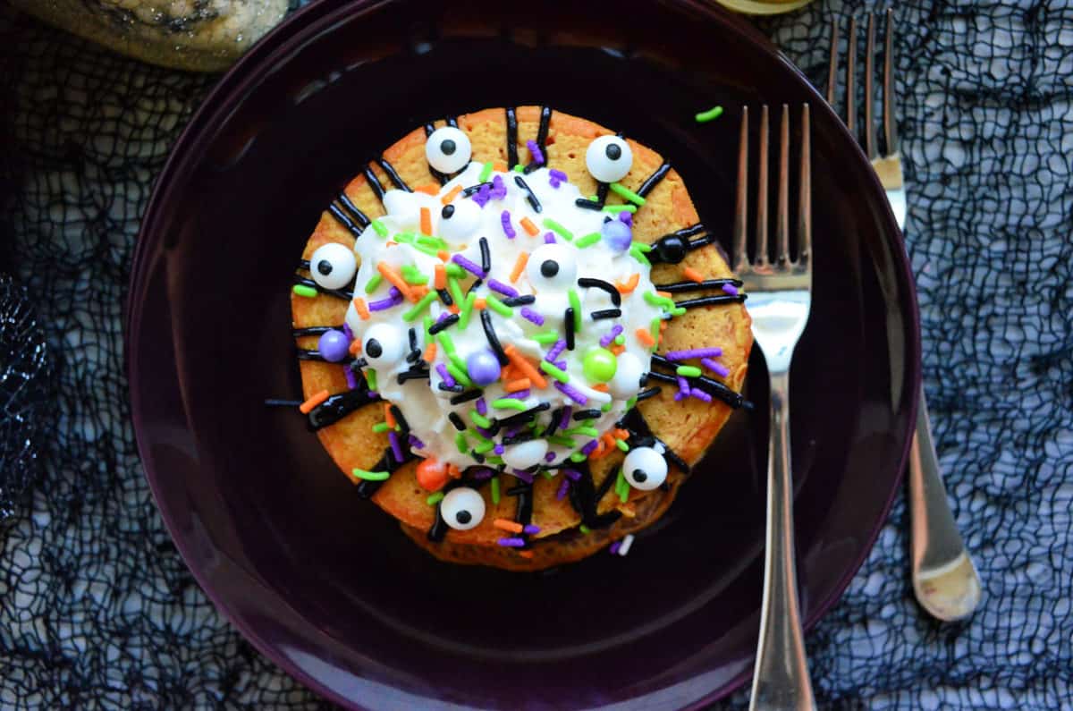 top view pancakes topped with whipped cream, sprinkles, and candy eyeballs on black plate.