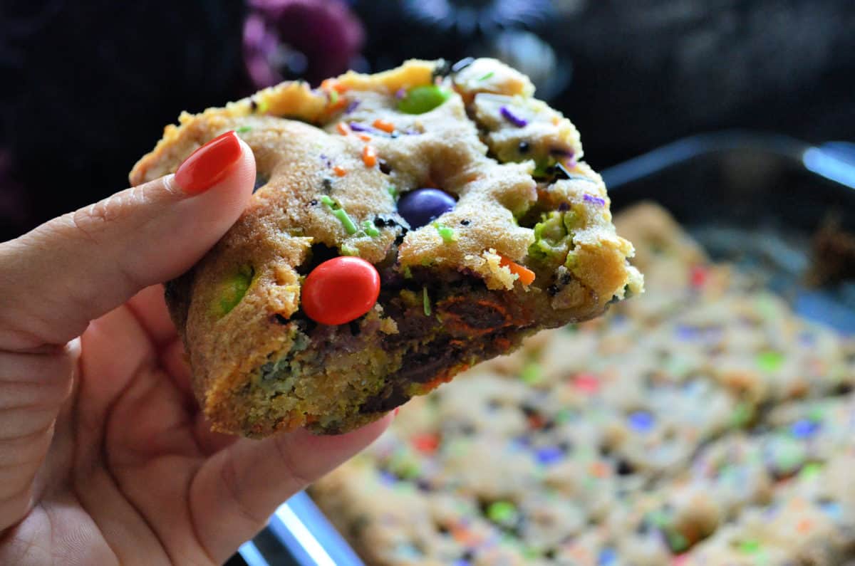 hand holding cookie bar square made with orange, purple, and green M&Ms and sprinkles up close.