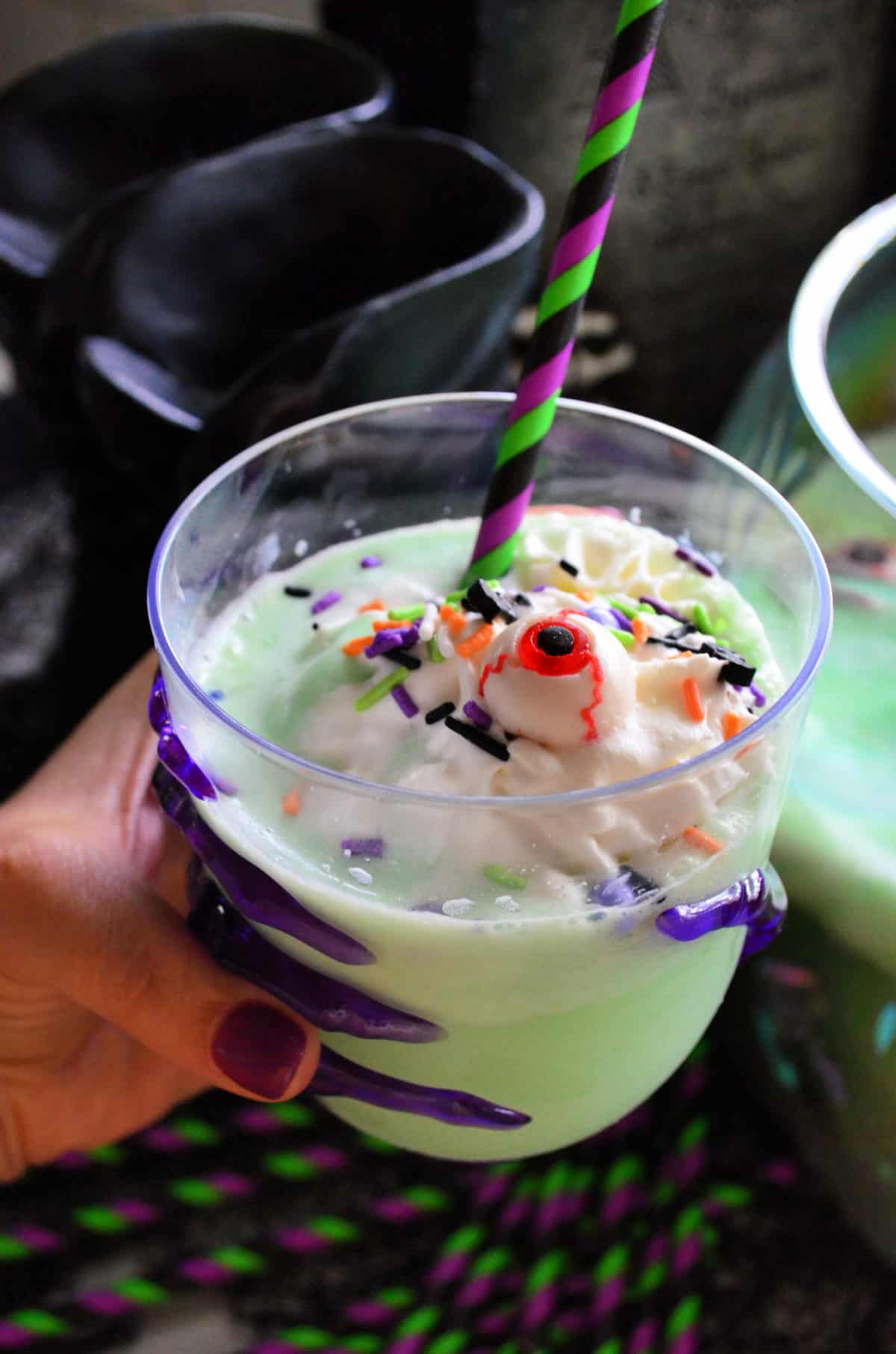 Close up of glass of creamy green drink topped with whipped cream, sprinkles, and candy monster eyeball.