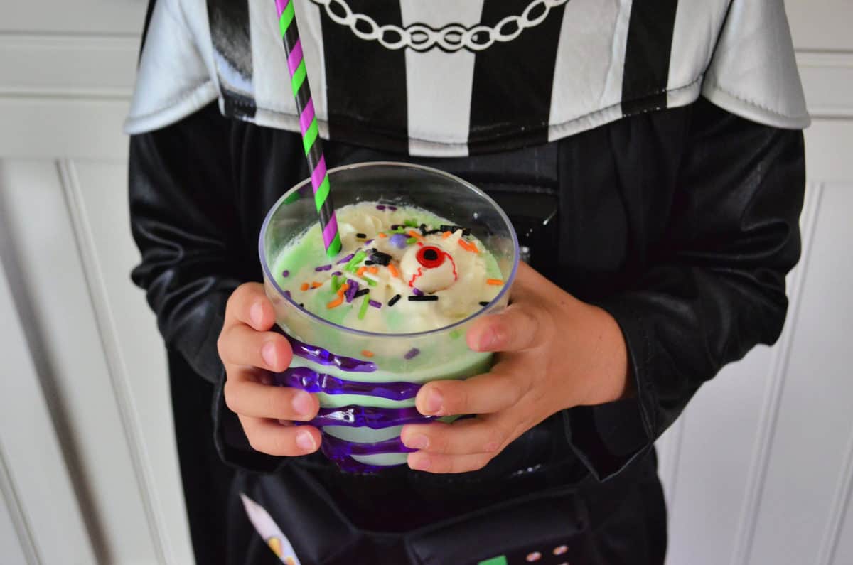 costumed kid holding cup of creamy green liquid topped with whipped cream, sprinkles, and candy eyeballs.