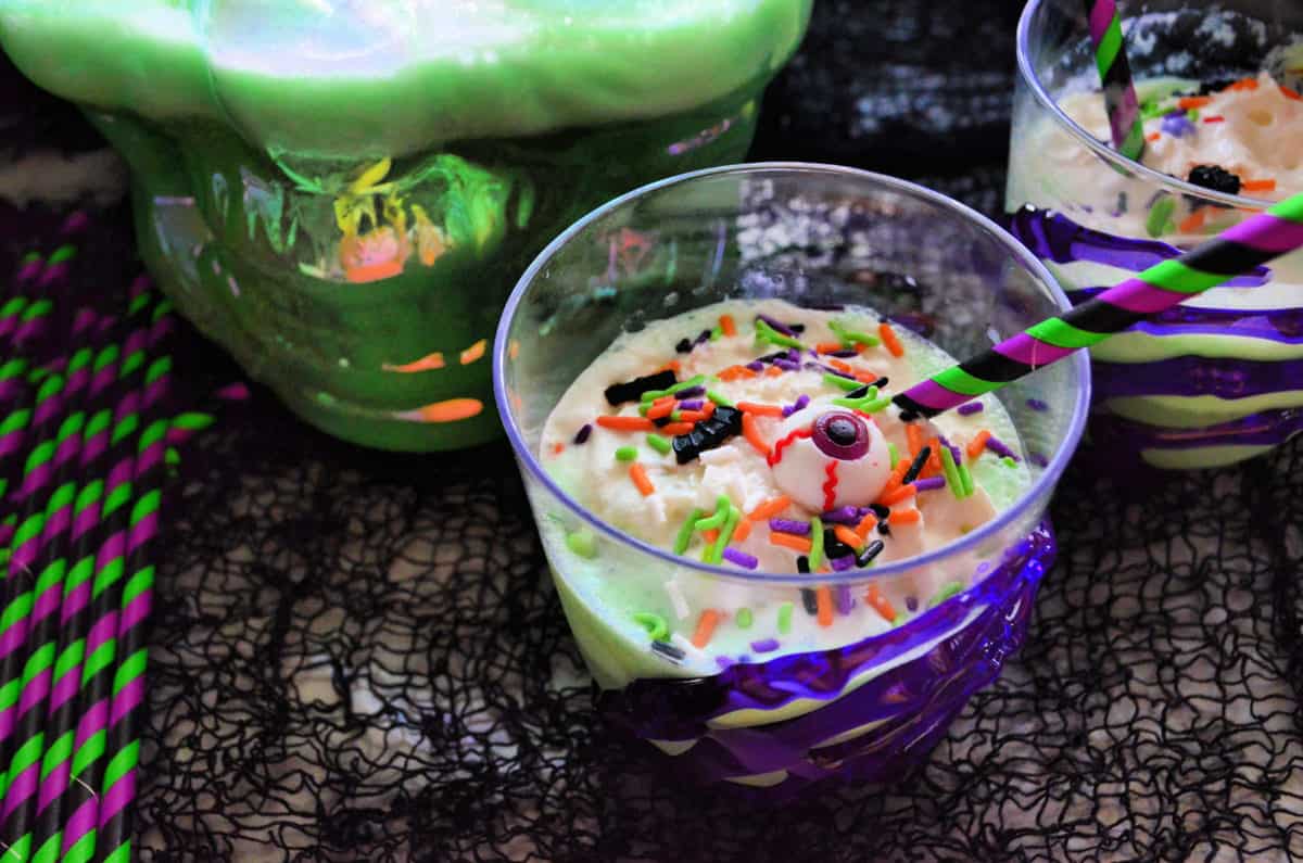 2 cups of creamy green liquid topped with whipped cream, sprinkles, and candy eyeballs on black gauze.