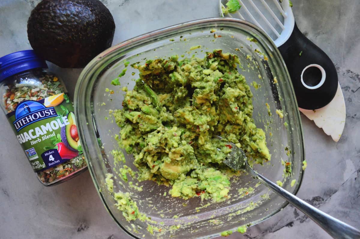 avocado being mashed with fork in glass bowl and mixed with lighthouse guacamole herb blend.