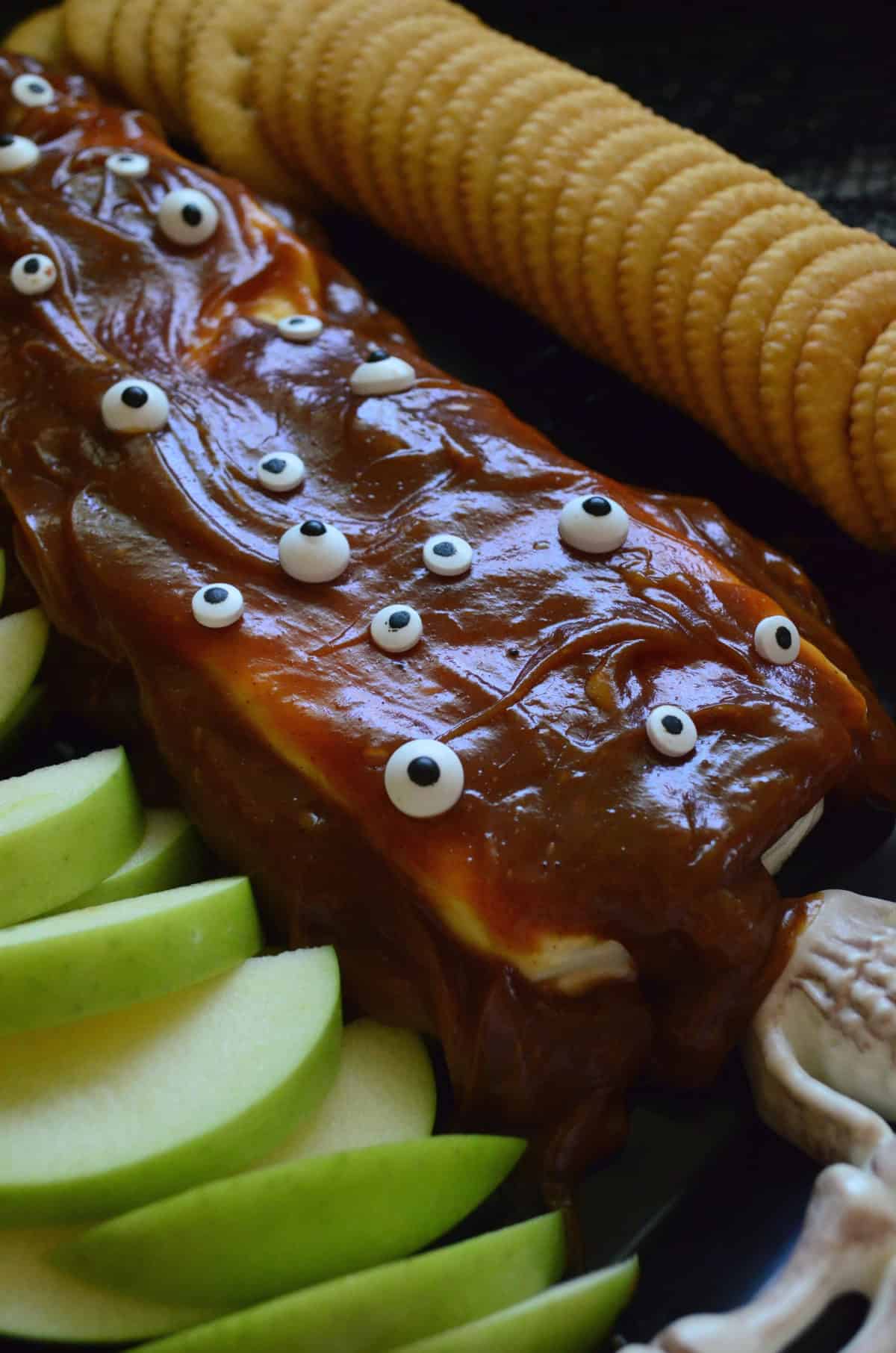 closeup of cream cheese covered in caramel and candy eyes served with apples and crackers.